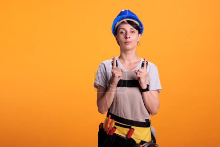 Photo for Portrait of woman builder pointing upwards with index fingers, showing above and indicating direction up. Posing in studio over yellow background and wearing hardhat with overalls. - Royalty Free Image