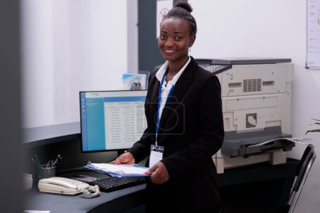Téléchargez les photos : Portrait de la réceptionniste en costume noir debout au comptoir de l'hôpital regardant les rendez-vous de visite de contrôle sur ordinateur. Employé d'accueil souriant vérifiant les documents médicaux dans le hall - en image libre de droit