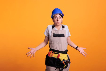 Photo for Construction worker doing i dont know sign and being clueless, acting uncertain and unsure in studio. Female contractor wearing hardhat and overalls, looking doubtful over background. - Royalty Free Image