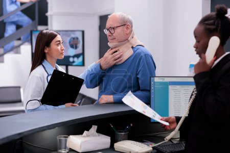 Photo for Old patient with cervical neck collar explaining fracture pain to doctor discussing health care treatment, standing at hospital registration desk. Man with medical foam waiting to attend consultation - Royalty Free Image