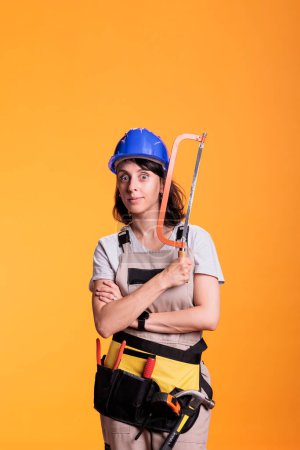 Téléchargez les photos : Femme charpentier professionnel utilisant la scie pour travailler sur le projet de rénovation, faire des travaux de rénovation en studio. Constructeur tenant une scie à main avec lame de rasoir, portant des combinaisons et un casque de protection. - en image libre de droit
