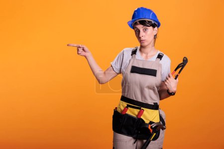 Photo for Woman builder indicating direction on left side of studio, posing with pair of pliers and pointing sideways. Female construction worker looking at camera and showing sides, yellow background. - Royalty Free Image