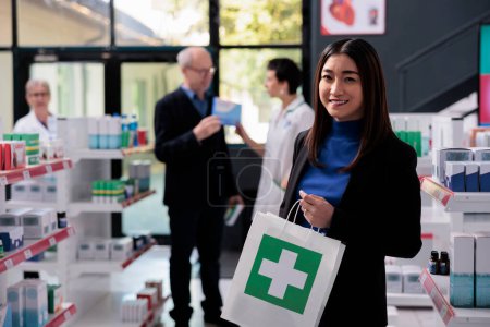 Photo for Smiling asian woman buying medication in drugstore and holding purchase portrait. Pharmacy store young buyer standing near shelf with medicament and carrying shopping paper bag - Royalty Free Image