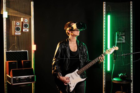 Photo for Woman performer playing heavy metal song using electric guitar while wearing virtual reality goggles during music session in sound studio. Performer with vr headset having grunge concert simultation - Royalty Free Image