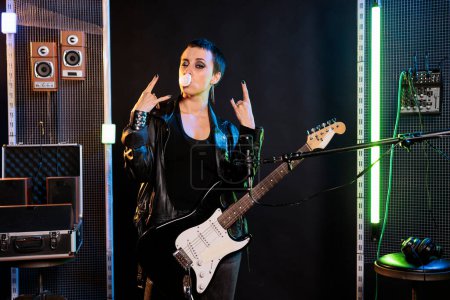 Photo for Woman performer blowing bubble gum while posing in studio doing rockstar gesture, preparing electric guitar before performing metal concert. Rebel musician playing for solo performance - Royalty Free Image