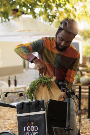 Photo for Young african american man putting bio organic produce in thermal bag, taking out order from farmers market and riding bike. Delivering locally grown fruits and vegetables to clients from farm. - Royalty Free Image