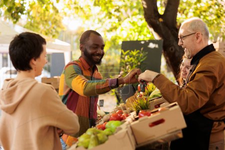 Photo for Elderly vendor giving apple sample to male customer, client trying out bio organic fresh fruits and vegetables. Senior seller small business owner selling organic eco products at farm stand. - Royalty Free Image