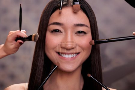 Téléchargez les photos : Brosses de maquillage autour sourire jeune asiatique mannequin visage. Belle femme heureuse obtenir professionnel nu naturel maquillage avec des cosmétiques décoratifs dans le salon de cosmétologie - en image libre de droit
