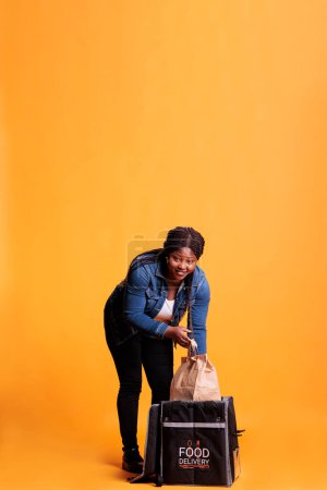 Foto de El empleado del restaurante saca una bolsa de papel de la mochila térmica que entrega el pedido de comida para llevar al cliente durante la hora del almuerzo. Deliverywoman de pie en el estudio con fondo amarillo - Imagen libre de derechos