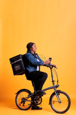 Téléchargez les photos : Pizzeria courier à vélo tout en livrant repas à emporter au client, vérifier adreess sur l'application de sortie de téléphone, debout sur fond jaune. Service à emporter et concept - en image libre de droit