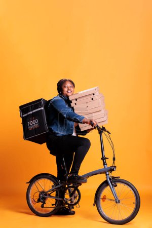 Téléchargez les photos : Travailleur du restaurant à vélo tout en portant pile de pizza livraison repas à emporter au client employé Pizzeria debout en studio avec fond jaune. Service alimentaire et transport - en image libre de droit