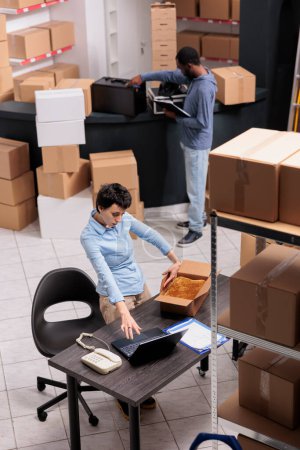 Photo for Top view of storehouse manager woman packing customer clothes order in cardboard box while checking shipping details on laptop computer. Team working in storehouse in delivery department. - Royalty Free Image