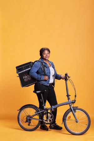 Téléchargez les photos : Courrier restaurant afro-américain debout à côté du vélo tenant sac à dos thermique tout en livrant de la nourriture à emporter au client pendant l'heure du déjeuner. Pizzeria employé portant repas à emporter - en image libre de droit