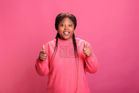 Foto de Sonriente afroamericano adulto joven haciendo pulgares hacia arriba signo mientras posando en el estudio sobre fondo rosa. Mujer alegre feliz con expresión facial alegre mostrando gesto de aprobación, persona haciendo como - Imagen libre de derechos