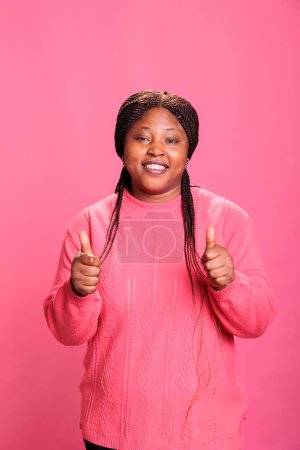 Téléchargez les photos : Modèle afro-américain souriant montrant pouce vers le haut signe debout sur fond rose en studio. Heureuse femme gaie avec expression joyeuse du visage donnant geste d'approbation, personne faisant comme signe - en image libre de droit
