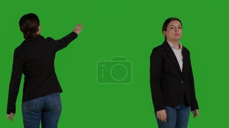 Photo for Close up of company employee raising hand and waving to call someone over, standing on isolated greenscreen backdrop. Young female worker asking and calling people to discuss, businesswoman. - Royalty Free Image
