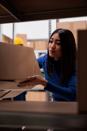 Photo for Goods supply chain manager doing inventory in warehouse while checking product box on shelf. Asian woman storehouse worker taking freight carton from rack in storage room - Royalty Free Image