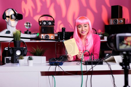 Photo for Smiling asian content creator holding notebook presenting school supplies in front of camera filming review in vlogging studio. Influencer with pink hair recording diary recommendation, live streaming - Royalty Free Image