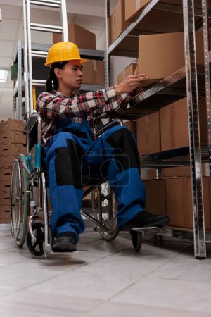 Téléchargez les photos : Jeune travailleur d'entrepôt asiatique en fauteuil roulant tout en faisant l'inventaire dans la salle d'entreposage. Responsable logistique avec handicap physique contrôle colis marchandises sur étagère dans l'entrepôt - en image libre de droit