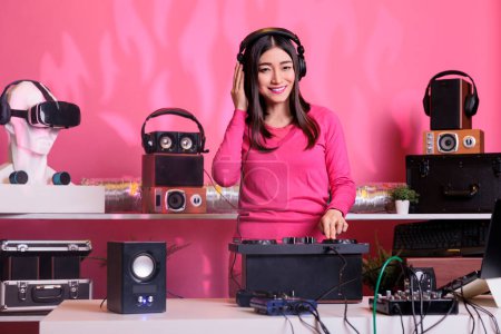 Photo for Smiling artist standing at dj table playing techno music at professional mixer console in studio over pink background. Asian musician performing electronic sound, having fun in club at night time - Royalty Free Image