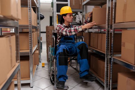 Photo for Young asian warehouse worker in wheelchair taking parcel from shelf. Storehouse order picker with physical disability searching cardboard box while working in storage room - Royalty Free Image