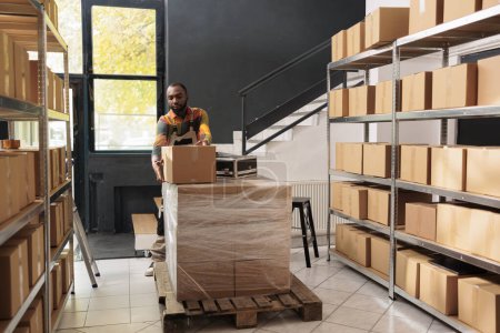 Photo for African american worker carrying cardboard box during warehouse inventory, preparing delivery for customers orders. Small business manager checking products doing quality control in storehouse - Royalty Free Image