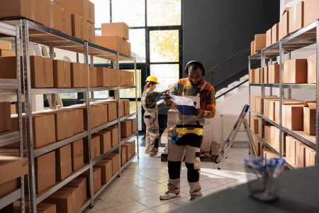 Photo for Cheerful employee having fun during storehouse inventory, listening music and dancing while analyzing merchandise checklist. Supervisor wearing protective overall preparing customers orders - Royalty Free Image