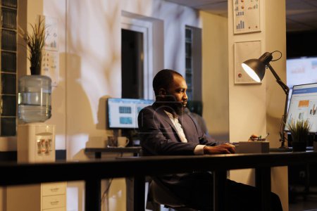 Téléchargez les photos : Homme d'affaires afro-américain debout à la table de bureau analysant le graphique financier sur ordinateur, travaillant à la stratégie de l'entreprise tard dans la nuit dans le bureau de démarrage. Plan d'investissement du gestionnaire exécutif - en image libre de droit