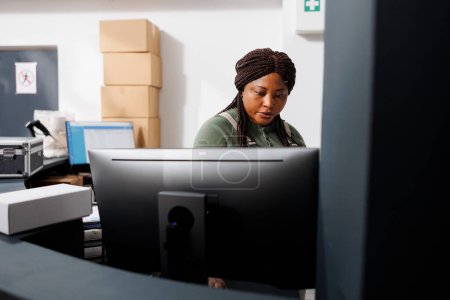 Photo for African american worker analyzing delivery details on computer, preparing customers packages for shipping in warehouse. Stockroom manager checking online orders during goods inventory - Royalty Free Image