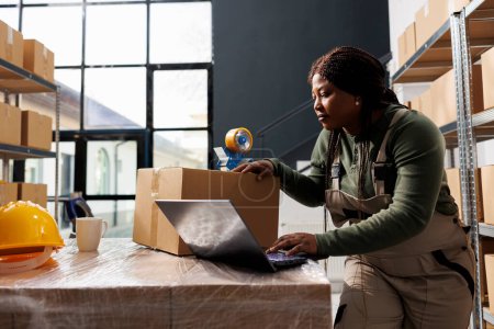 Téléchargez les photos : Travailleur afro-américain vérifier la logistique de l'entrepôt sur ordinateur portable, préparer les commandes des clients pour la livraison. Employé de la salle de stockage travaillant à l'inventaire des marchandises dans l'entrepôt - en image libre de droit