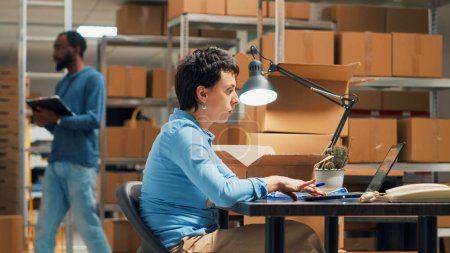 Photo for Female employee working on shipping stock from storehouse shelves, using products and laptop. Small business owner planning distribution shipment, looking at cardboard boxes. Handhed shot. - Royalty Free Image