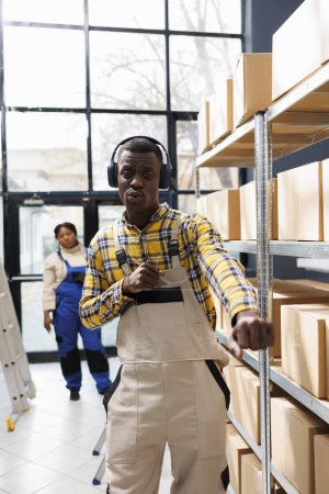 Photo for Warehouse package handler dancing at work and looking at camera. African american storehouse worker listening to playlist in headphones, singing along and making arms movements portrait - Royalty Free Image