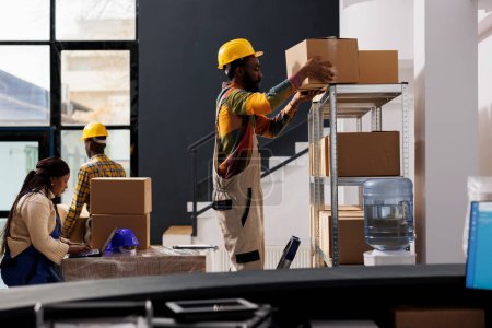 Photo for African american warehouse managers team picking buyer order and choosing cardboard box for packing. Woman checking goods list on laptop and man standing on ladder while taking parcel from shelf - Royalty Free Image