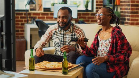 Photo for Cheerful man and woman eating slices of pizza at home, being relaxed together watching movie on television. Young people in relationship enjoying delivery meal and beer bottles. - Royalty Free Image