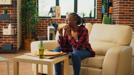 Photo for African american woman watching tv and eating pizza, laughing at comedy movie in living room. Young person serving fast food delivery meal and binge watching favorite tv show. - Royalty Free Image