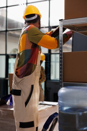 Téléchargez les photos : Un Afro-Américain debout sur une échelle et fouillant un colis sur une étagère dans un entrepôt industriel. Ouvrier d'entrepôt portant le chapeau dur prenant la boîte en carton pour l'emballage de commande de client dans l'entrepôt - en image libre de droit