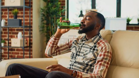 Téléchargez les photos : African american person eating burger and fries on sofa, having dinner with fast food and bottle of alcohol at home. Young man feeling happy watching action movie or show, having fun. - en image libre de droit