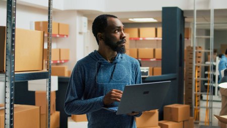 Photo for Male storage room worker using laptop to write products data, analyzing racks filled with cardboard packages for stock inventory. Young man planning logistics and merchandise management. - Royalty Free Image