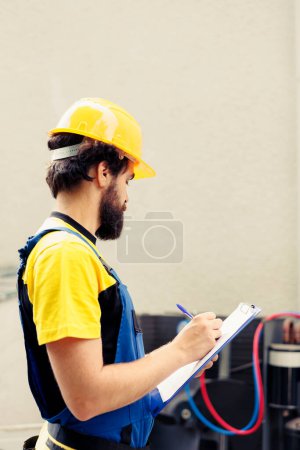 Photo for Experienced technician commissioned for outdoor air conditioner annual maintenance, writing report on clipboard. Skilled engineer doing hvac system inspection, looking for faulty components - Royalty Free Image