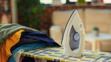 Foto de Iron tool placed on ironing board with pile of washed clothing, electric appliances used to do housekeeping work. Domestic chores with iron to smooth out creases on laundry. Close up. - Imagen libre de derechos