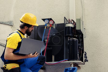 Photo for Licensed technician commissioned for outside hvac system annual maintenance, looking for refrigerant leaks. Certified expert doing air conditioner inspection, imputing data on laptop - Royalty Free Image