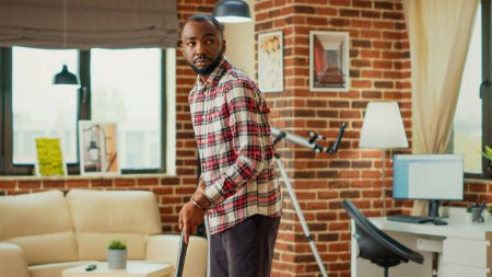 Foto de African american adult doing spring cleaning session at home, using vacuum cleaner to tidy up living room. Young man vacuuming wooden apartment floors, using all purpose cleaner. - Imagen libre de derechos