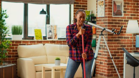 Téléchargez les photos : African american housewife listening to music and mopping apartment floors, using mop and washing solution. Young happy woman dancing and singing, having fun with spring cleaning. - en image libre de droit