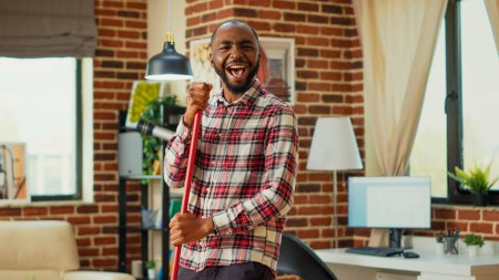 Photo for Modern happy boyfriend having fun with music while he uses mop to wash floors, mopping in apartment. Young male adult dancing and singing in living room, spring cleaning enjoyment. - Royalty Free Image