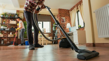 Photo for African american man vacuuming floors in living room, cleaning apartment with girlfriend. Young adult using vacuum cleaner and woman wiping shelves with all purpose cleaner. Tripod shot. - Royalty Free Image