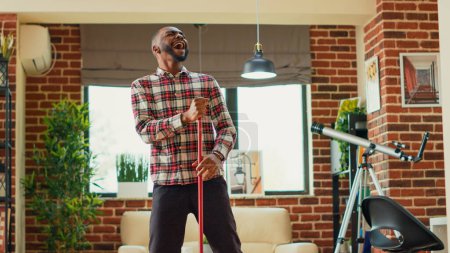 Foto de Afroamericano adulto fregando salón pisos de madera y bailando, actuando tonto con movimientos de baile en casa. Hombre feliz haciendo limpieza de primavera con fregona, divirtiéndose y sonriendo. Disparo de mano. - Imagen libre de derechos