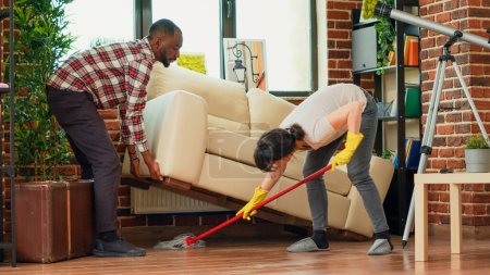 Photo for Diverse couple washing wooden tiles in living room, boyfriend helping girlfriend to clean floors under couch. Young housewife mopping apartment ground with cleaner. Handheld shot. - Royalty Free Image