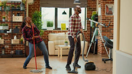 Téléchargez les photos : African american partners listening to music and washing floors in living room, doing silly funny dance moves. Cheerful people doing spring cleaning and dancing in apartment, mopping. - en image libre de droit