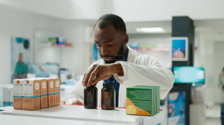 Téléchargez les photos : Male specialist putting stock of medicaments and drugs on pharmacy shelves, putting boxes of pills and vitamins to help clients. Young adult working with pharmaceutical products, healthcare. - en image libre de droit