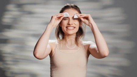 Téléchargez les photos : Beauty model acting silly with cotton pads over eyes, playing around in front of camera. Cheerful glowing woman having fun laughing in studio, posing with luminous radiant skin. - en image libre de droit